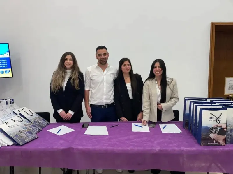 נתנאל לומר ושלוש עמיתותיו עומדים מאחורי שולחן עם מסמכים חתומים, מביטים למצלמה ומחייכים